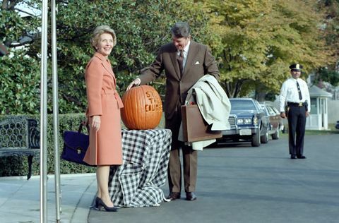 na této fotografii prezident Ronald Reagan obdivuje dýni vyřezanou se sloganem „zůstaň v kurzu“, prezident Reagan použil tento slogan během 1982 průběžné volby první dáma Nancy Reaganová pózuje vedle dýně, ti dva stojí před jižním portikem, vchodem do bílé Dům