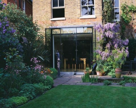 Pohled na trávník a zadní část domu s obdélníkovým bazénem, ​​vistárií, prosklenou kuchyní a sochou Heleny Sinclairové