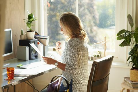 Žena, pití kávy a čtení papírování u stolu v slunné domácí kanceláři.