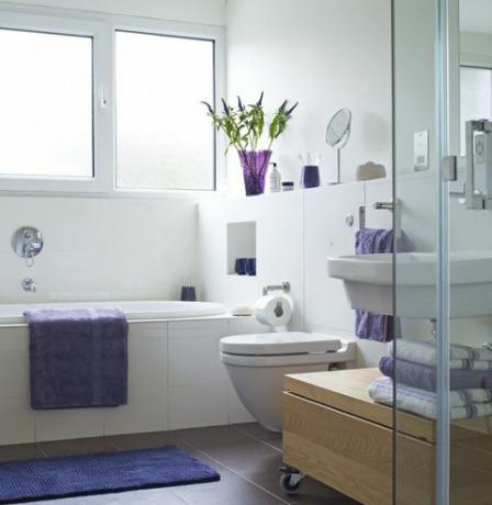 Světle osvětlená koupelna s fialovým ručníkem na boku vany a složené ručníky poblíž sprchy