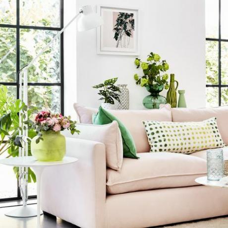 růžová pohovka v bílé místnosti se zelenými rostlinami kolem a zelenými vzorovanými polštáři